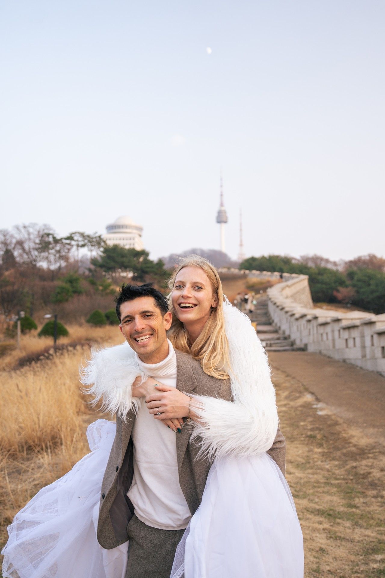 A couple laughs and embraces while taking outdoor photos along a stone wall on a sunny autumn day.