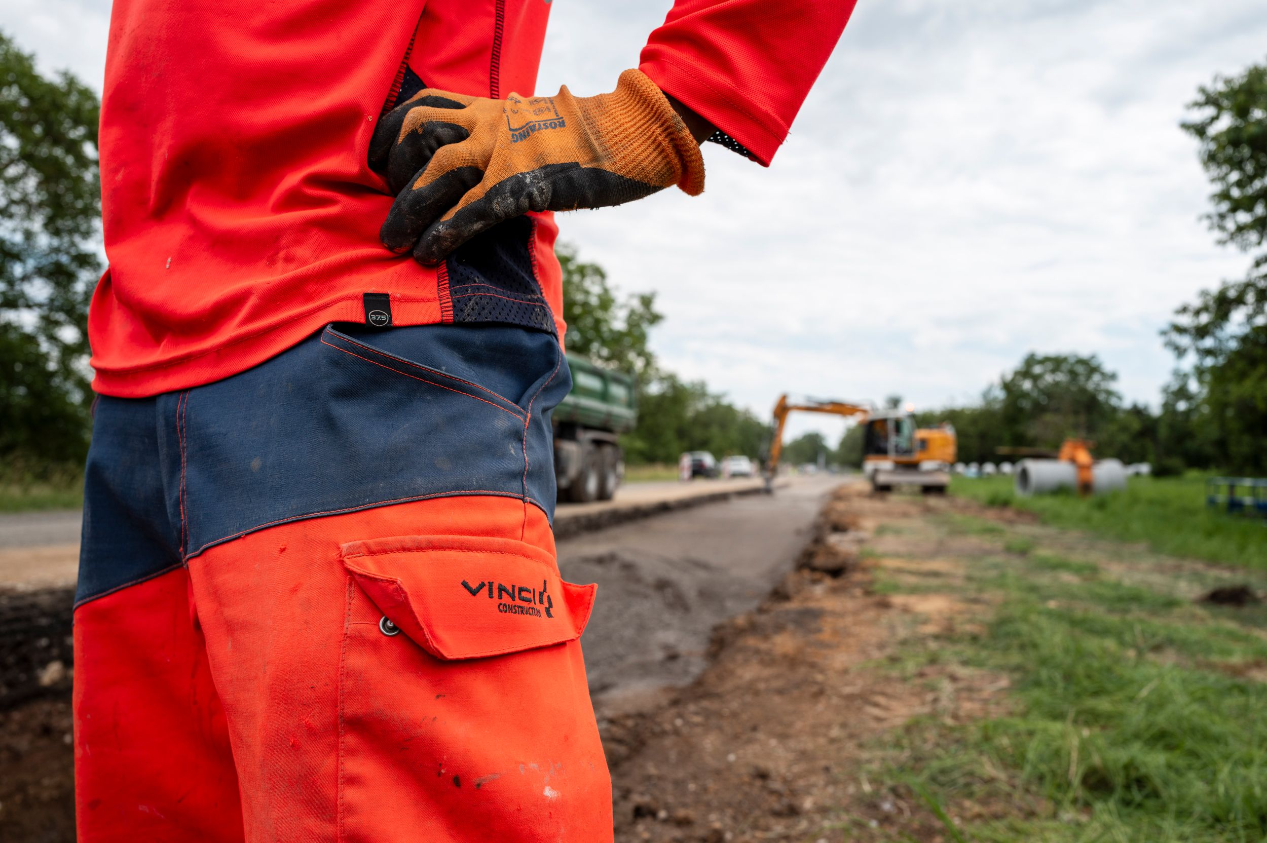 Construction worker in red safety clothing and work gloves stands on a dirt road with heavy equipment in background.
