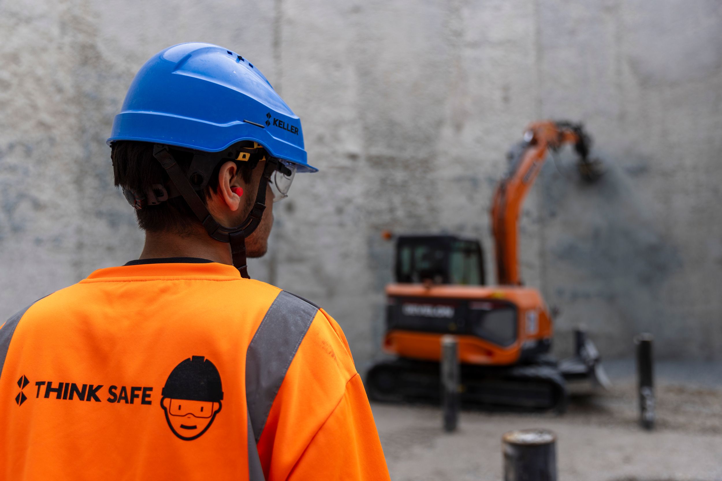 Construction worker in orange safety vest and blue helmet watches excavator operate at a job site.