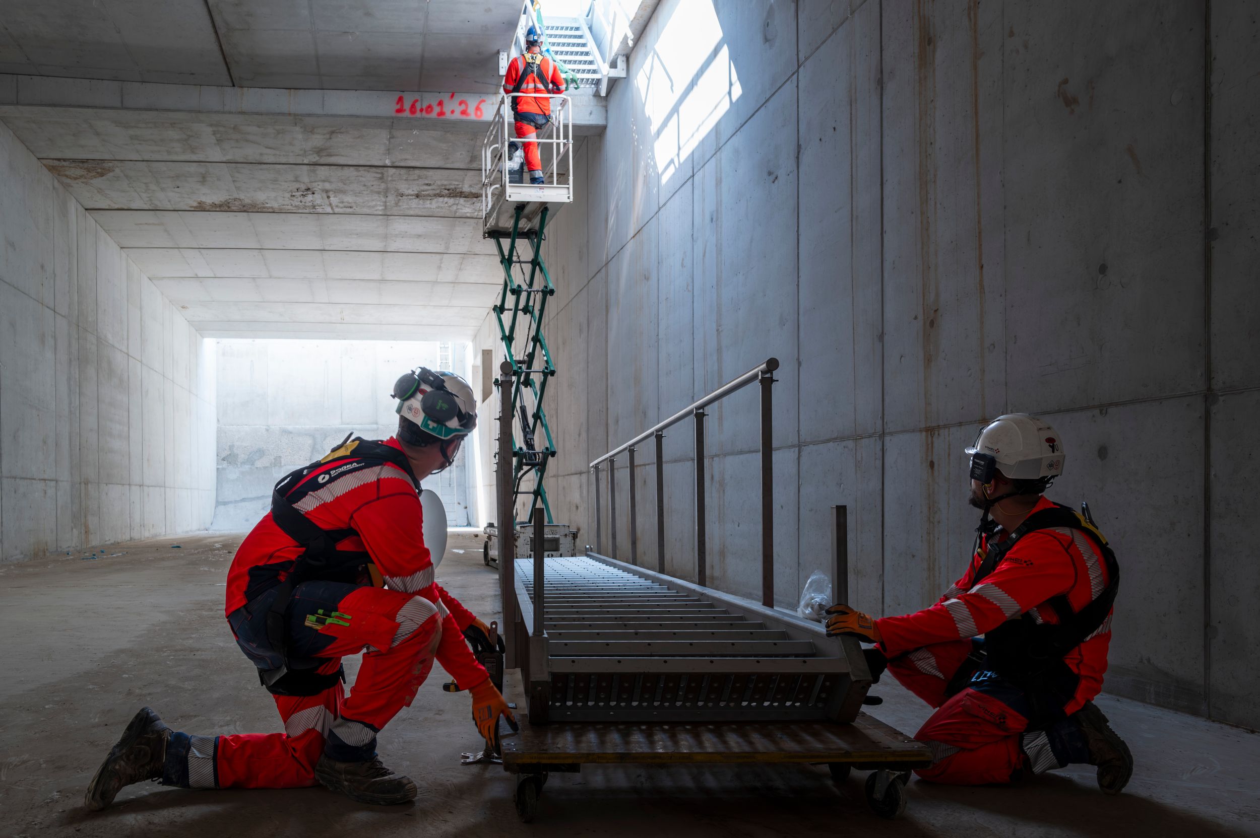 Workers in red safety gear assemble equipment inside a concrete tunnel, with a colleague on a scissor lift in the background.