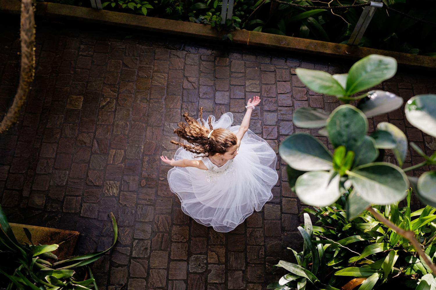 A bride in a flowing white wedding dress twirls on a brick pathway surrounded by lush greenery and gardens.