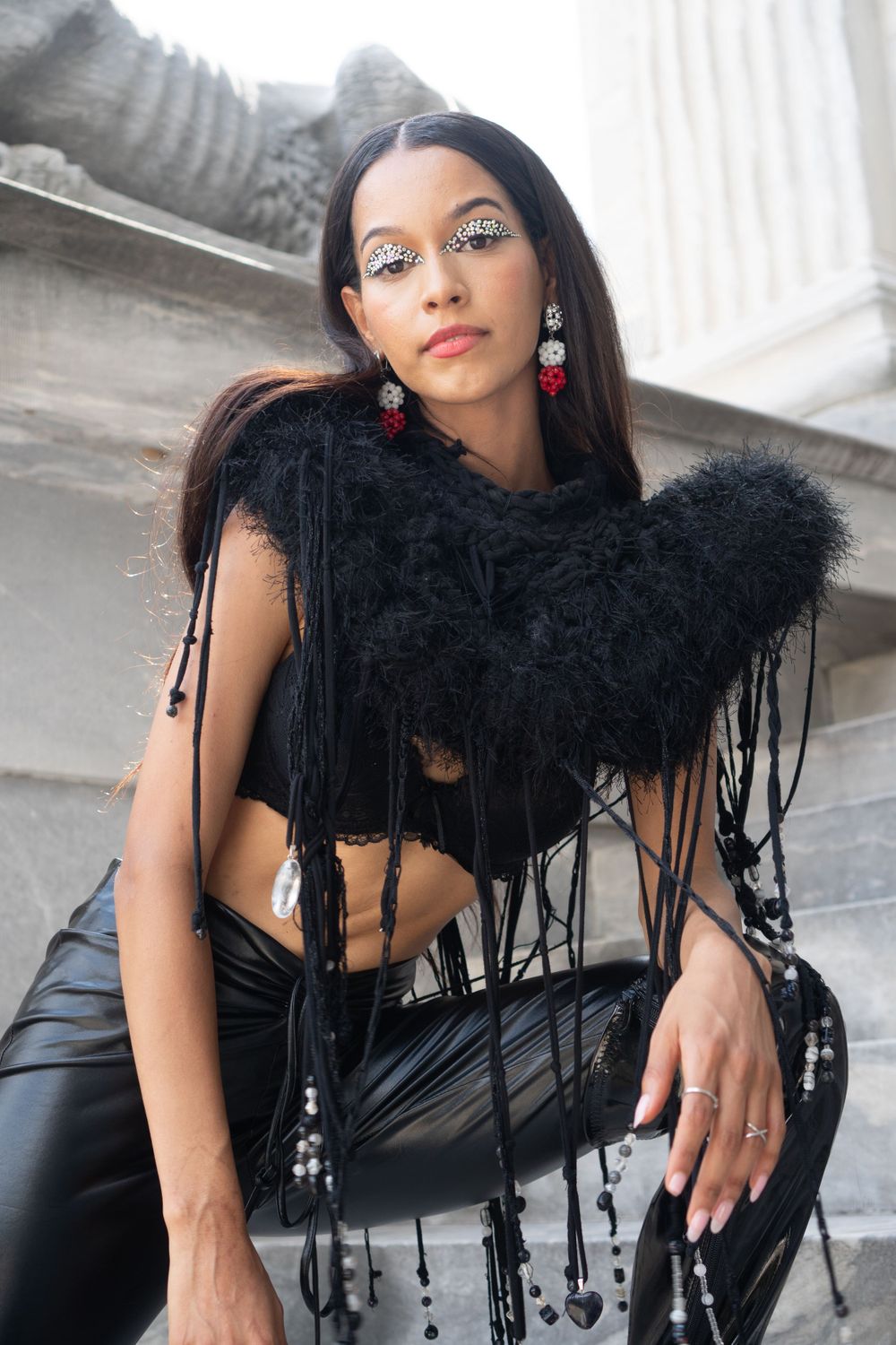 Fashion model in edgy black outfit with feathers and red earrings against stone columns in Philadelphia.