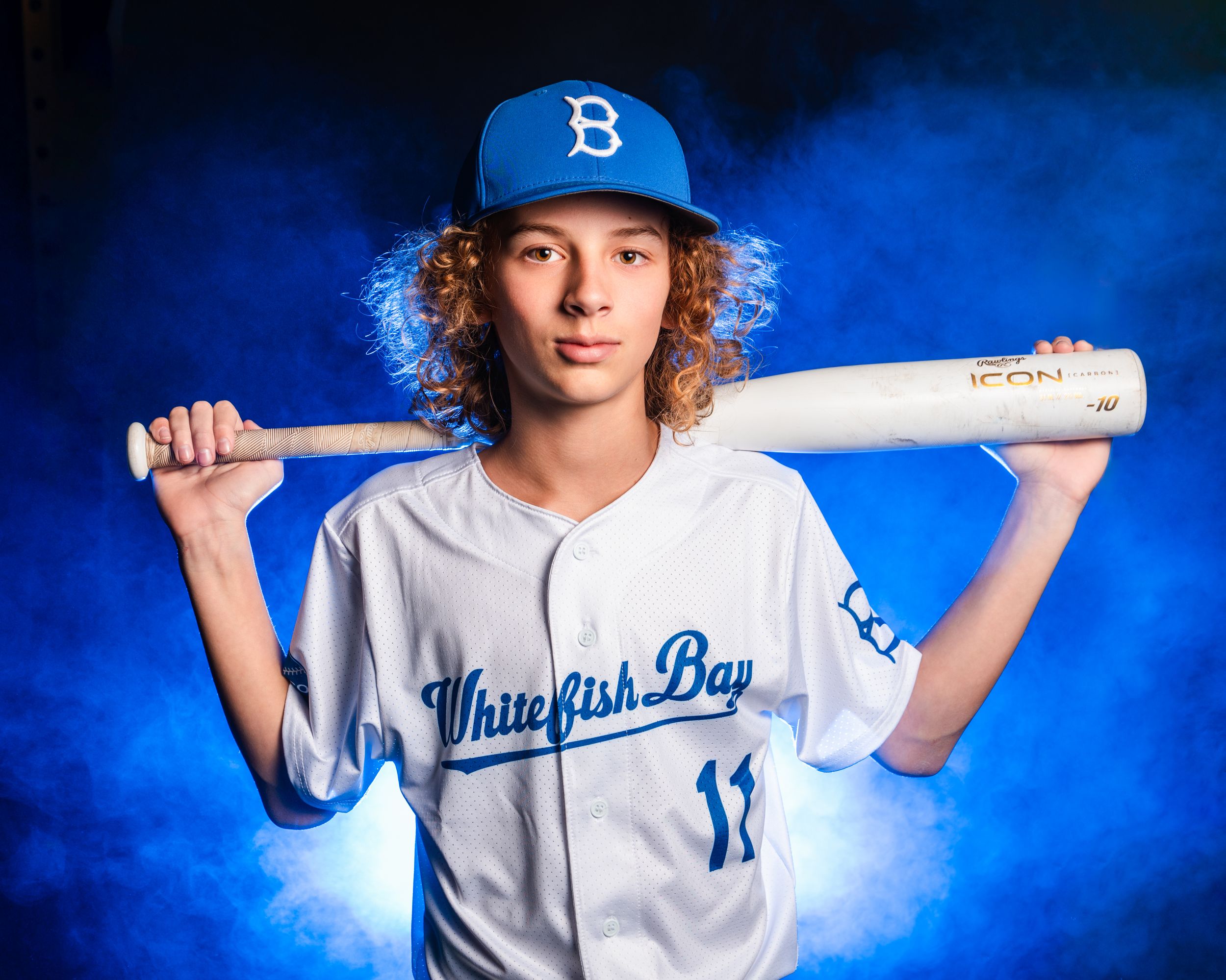 Baseball player in white Whippany Bay uniform holds wooden bat over shoulder against blue backdrop.