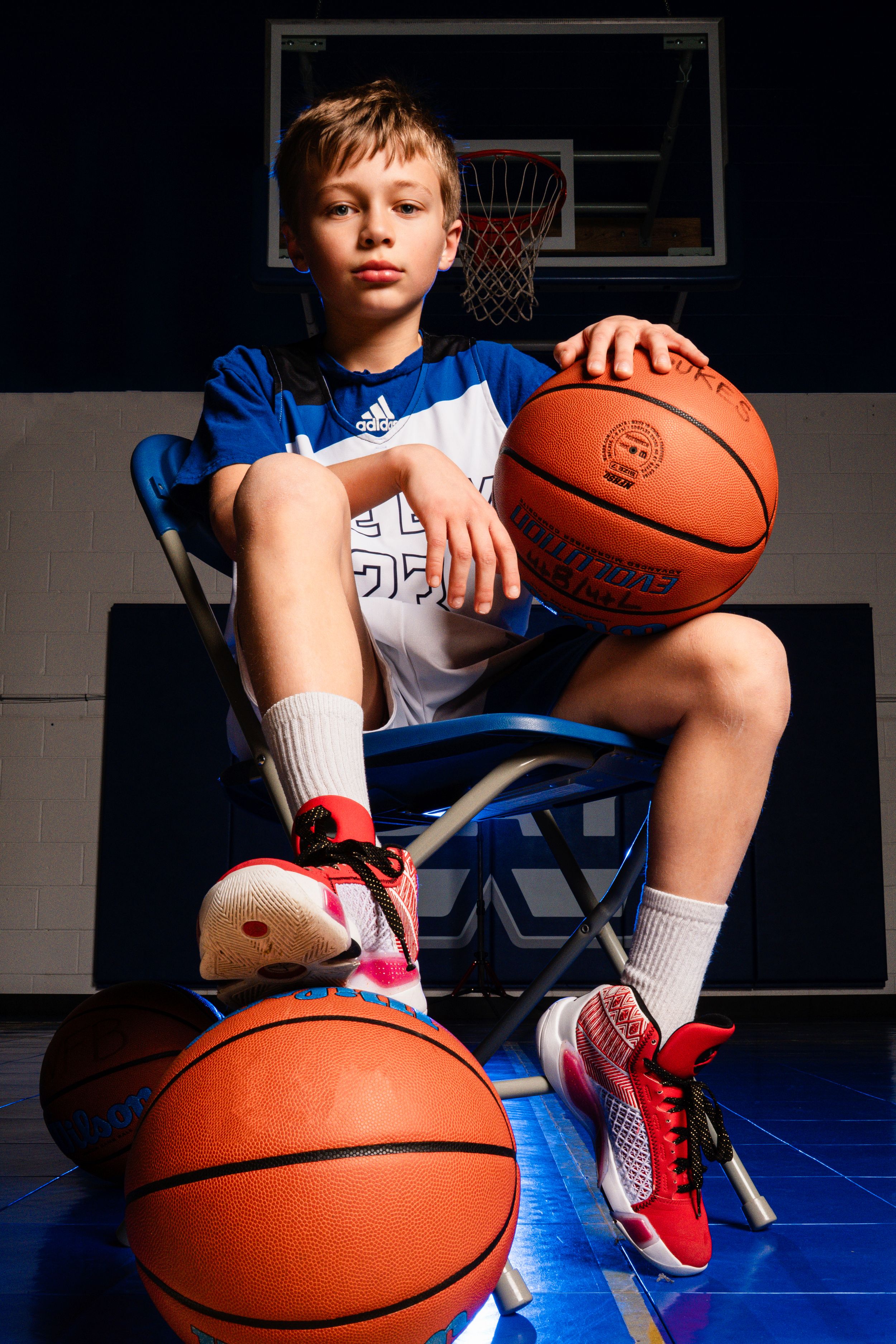 Young basketball player in blue jersey poses with basketballs against backboard backdrop on blue court.