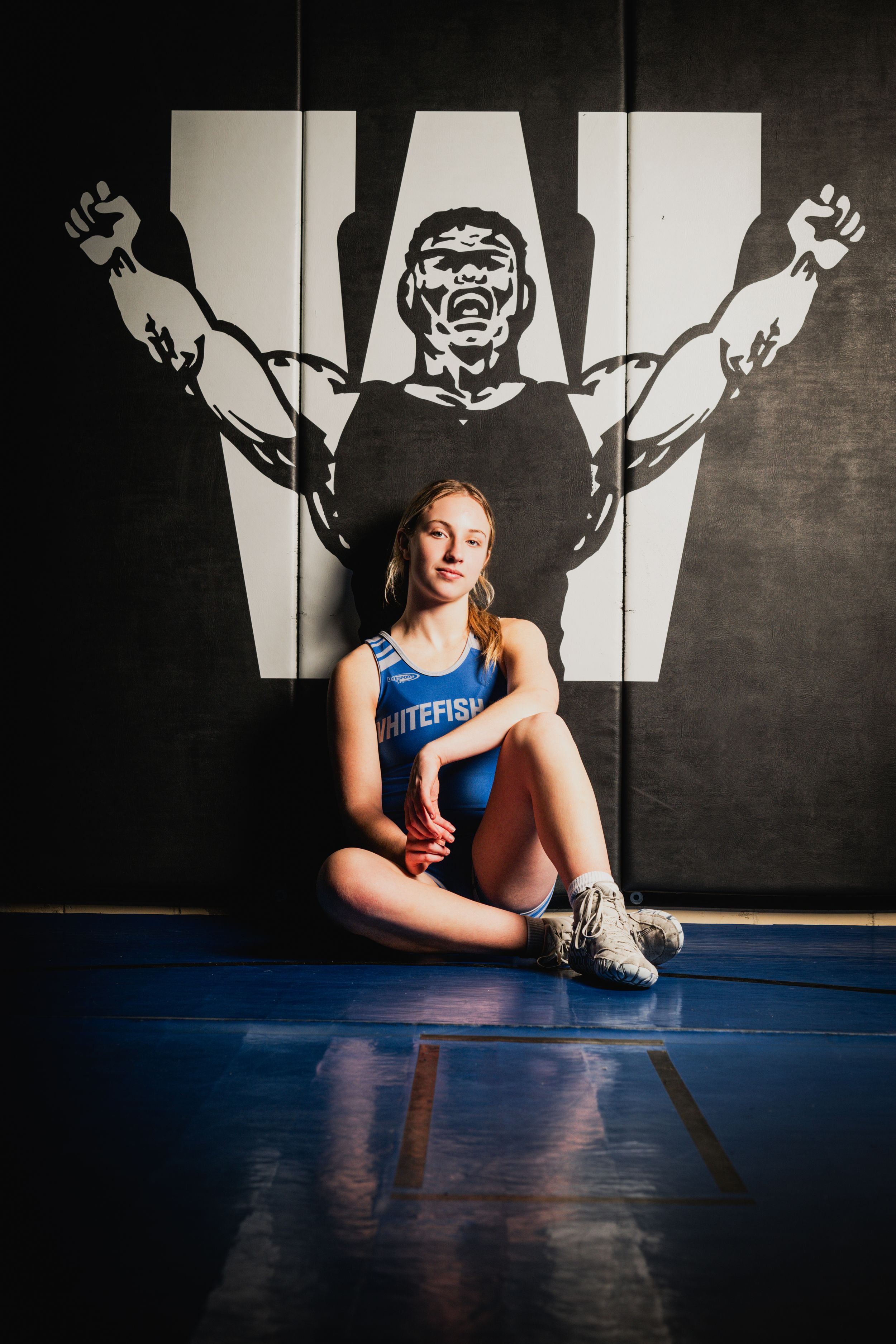 Athlete sitting on floor against wall with dramatic muscular shadow silhouette backdrop in gym setting.