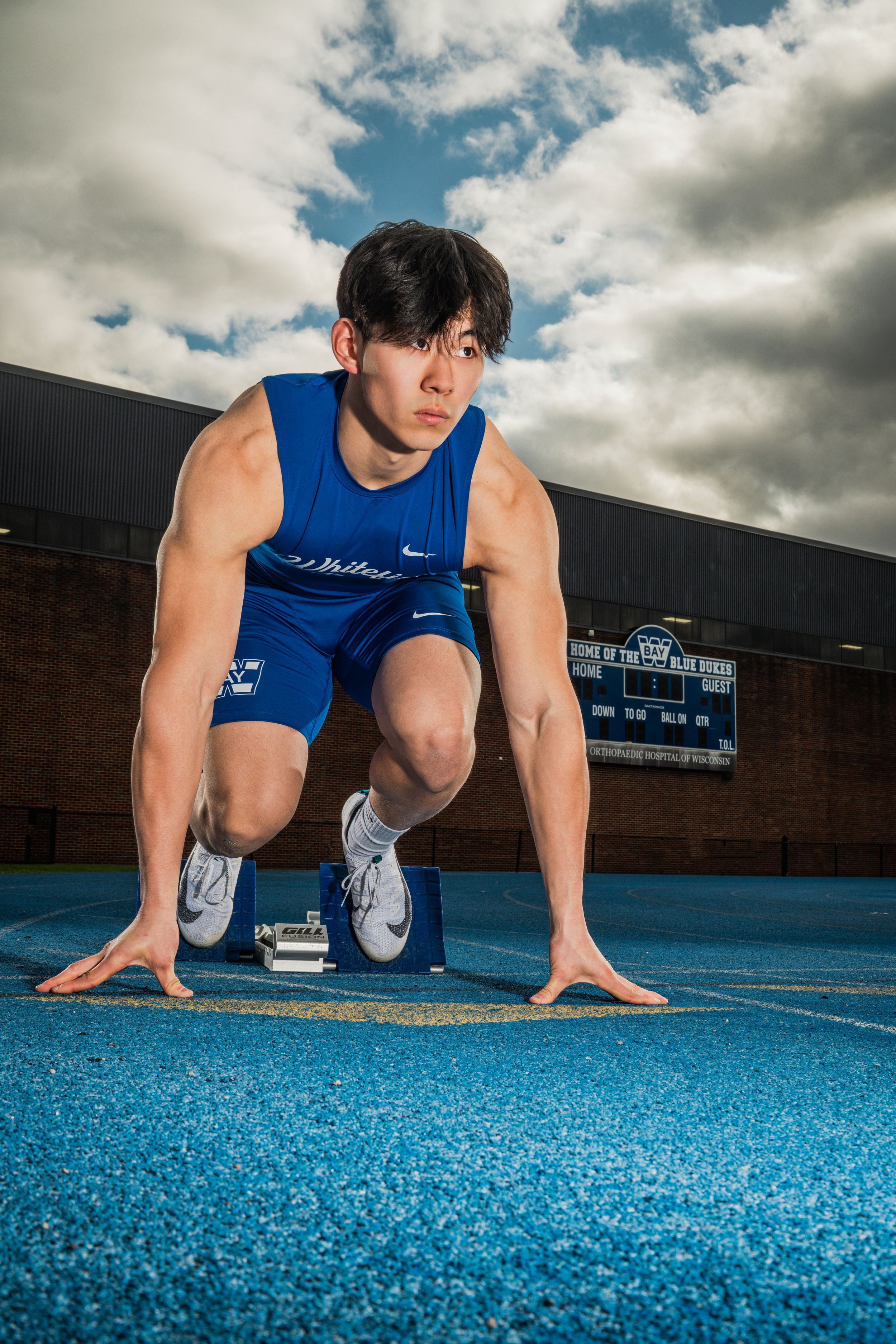 Athlete in blue athletic wear doing sprinting drills on a blue track surface under cloudy skies.