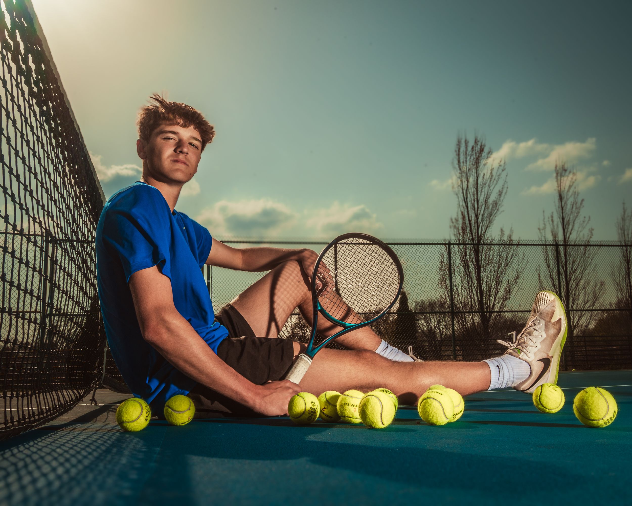 Tennis player sitting on court with tennis balls and racket at sunset with dramatic lighting and chain link fence.