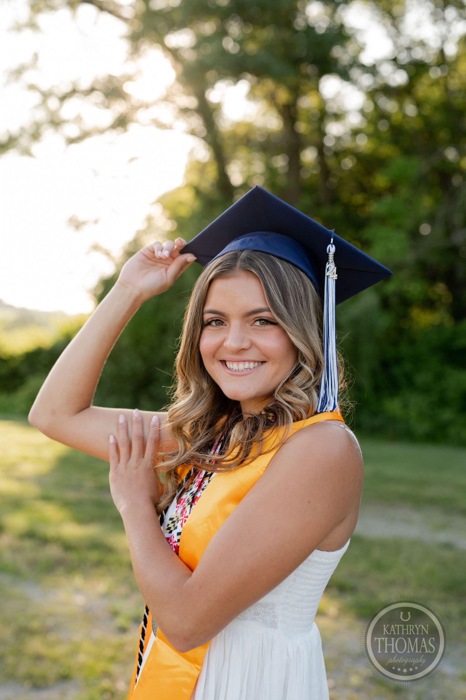 Graduate in white dress and academic regalia poses with hand on cap in evening light.