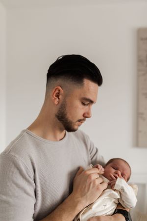 A tender moment shows a young parent in a light gray sweater tenderly holding a sleeping newborn swaddled in white.