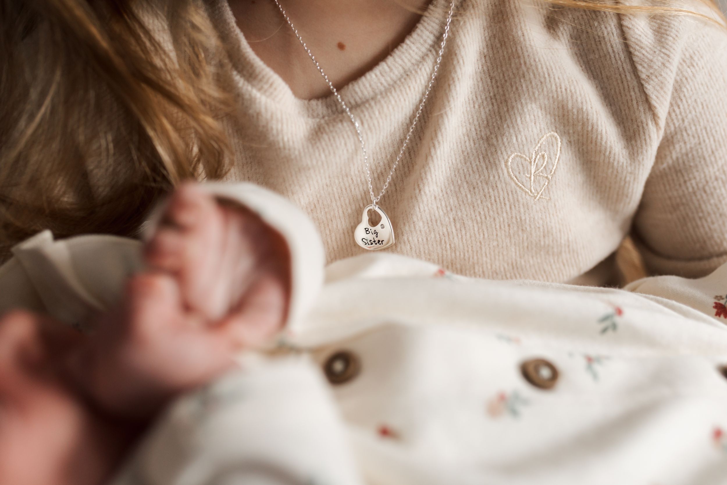 A delicate silver heart pendant necklace worn with a cream colored sweater in soft natural lighting.