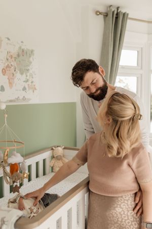 A couple stands lovingly at a white crib in a nursery with a world map on the wall and soft green accents.