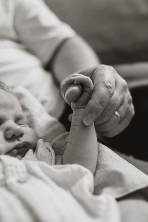 A black and white close-up photograph shows tiny newborn baby fingers grasping an adult's hand.