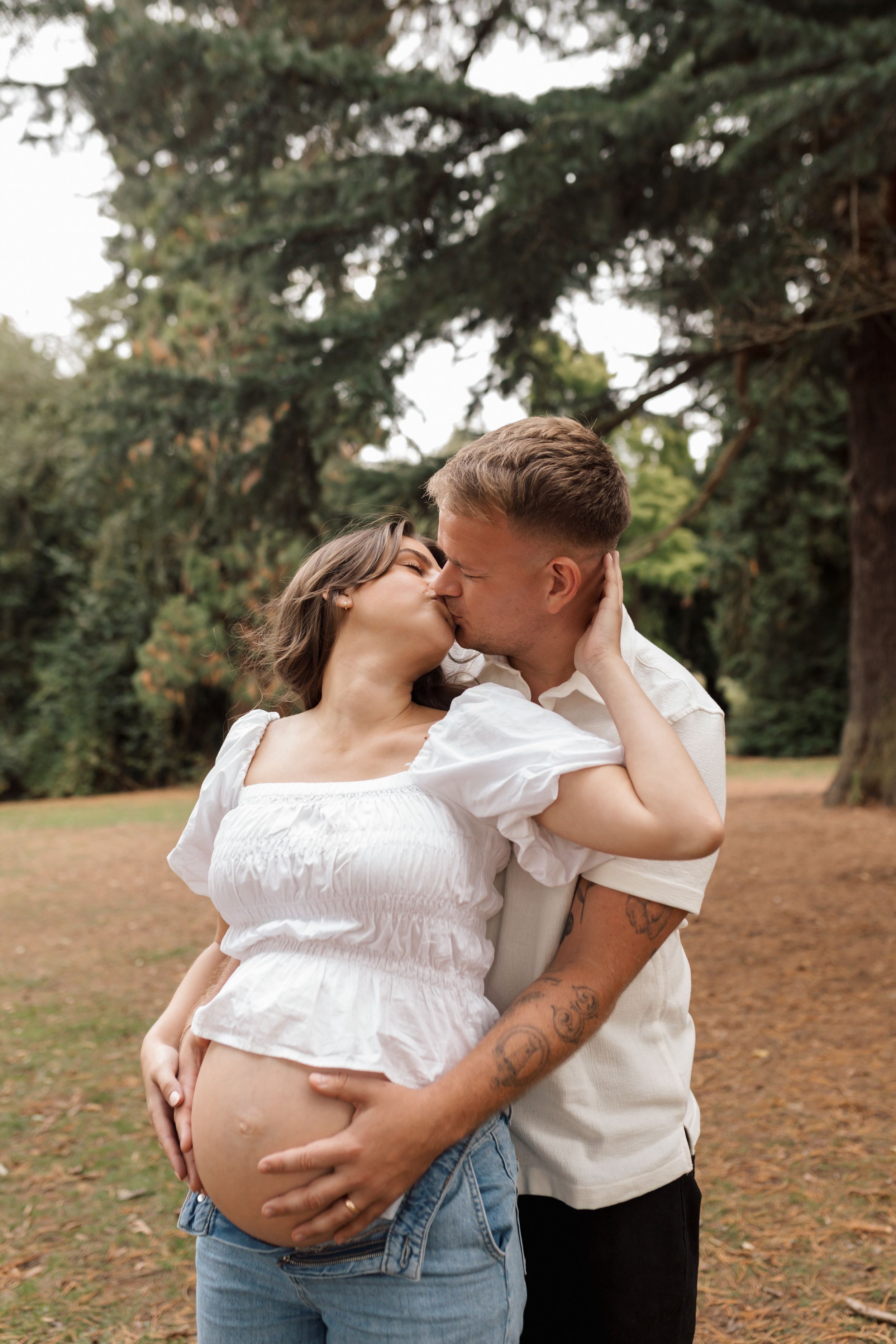 Romantic couple in white tops and jeans embrace and kiss in an outdoor park setting with evergreen trees.