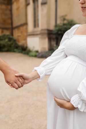 A pregnant woman in a white dress holds hands with someone while cradling her belly outdoors.