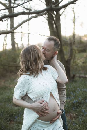 Intimate maternity photo outdoors with a tender embrace and kiss in a wooded setting at golden hour.