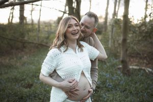 Couple shares tender moment in wooded setting during maternity photoshoot, woman wearing white lace dress.