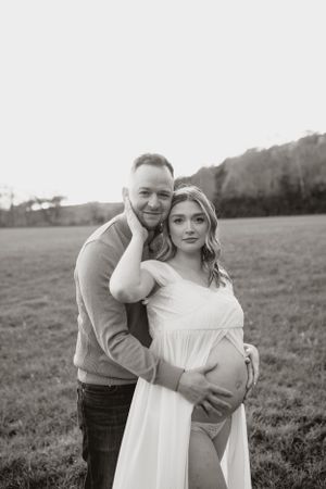 A romantic black and white portrait of an expecting couple embracing in a field at sunset.