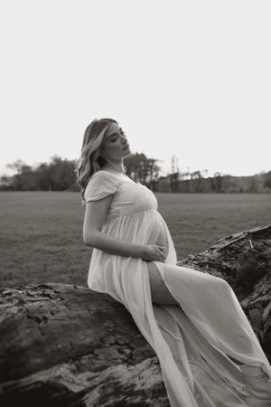Black and white maternity photo of a woman in a flowing white dress posing on rocks in a field during sunset.