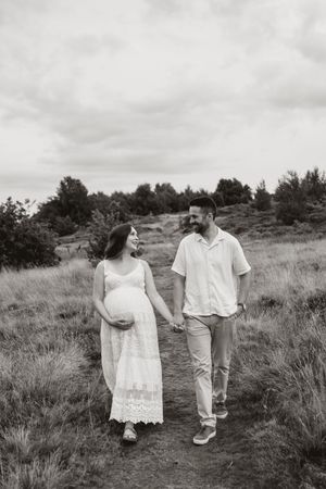 A couple in white clothing walks together through a grassy meadow on a cloudy day.