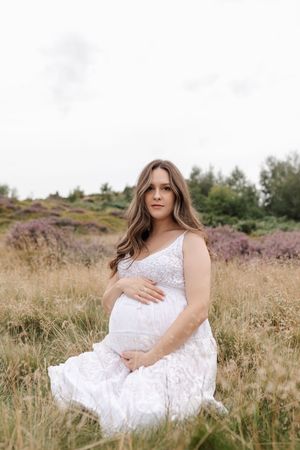 A woman in a white lace dress poses gracefully in a field of tall grass and wildflowers during a maternity photoshoot.