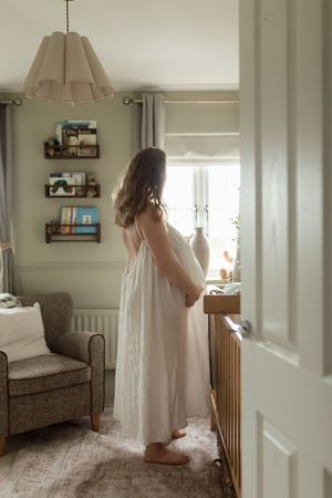 Expecting mother in white nightgown stands by window in cozy nursery with floating shelves and vintage armchair.