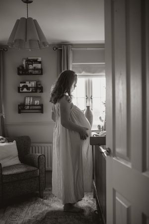 A silhouetted pregnant figure in a white dress stands by a window in a cozy nursery room with floating shelves and soft lighting.
