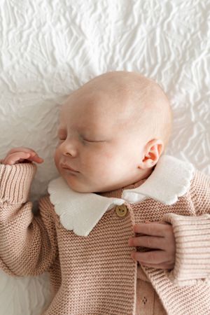 Sleeping infant wearing soft pink knit sweater with white peter pan collar rests on white textured bedding.