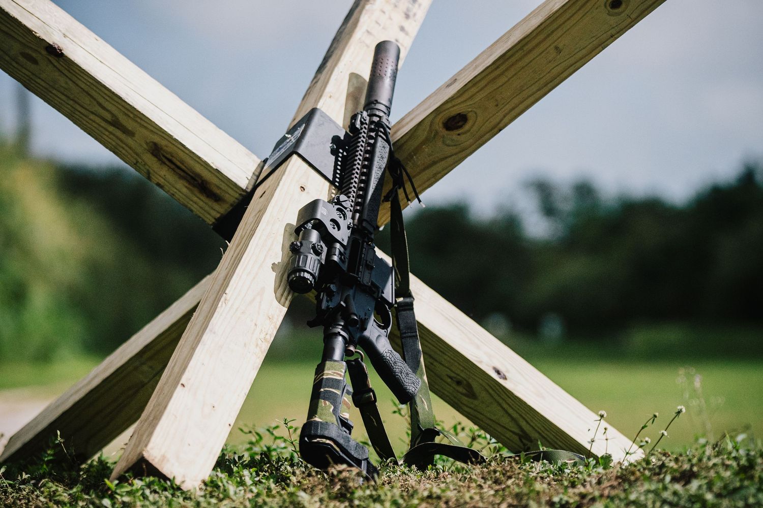 A tactical rifle leans against wooden cross beams in an outdoor setting.