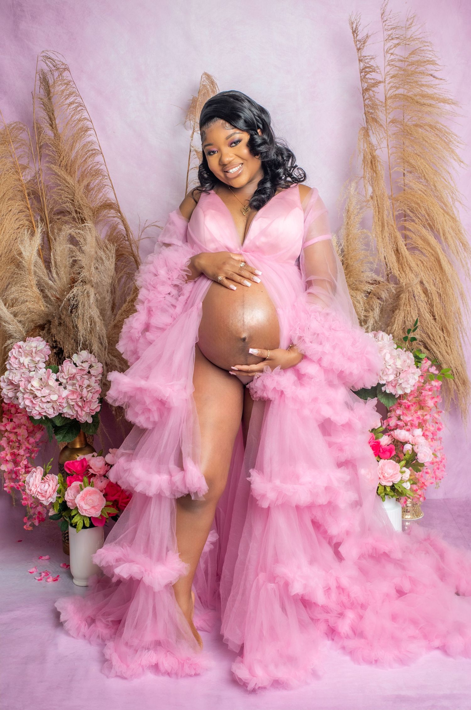 Maternity photoshoot with flowing pink tulle robe against floral backdrop with pampas grass and roses.