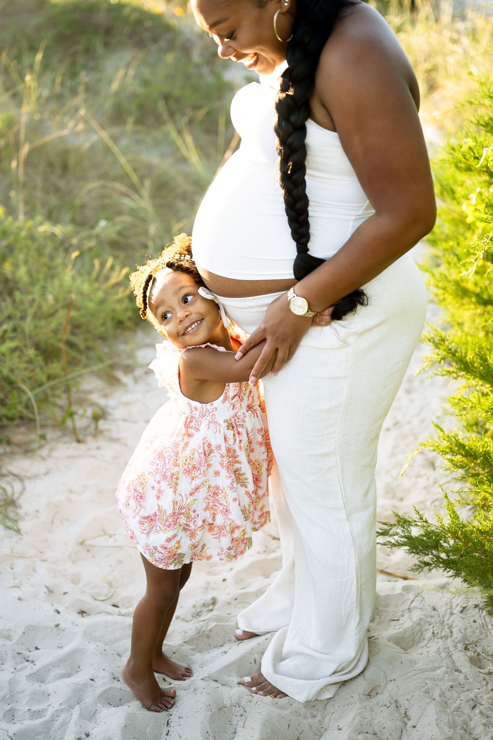 A pregnant woman in white attire stands on a beach with a young child in a floral dress during a sunny day.