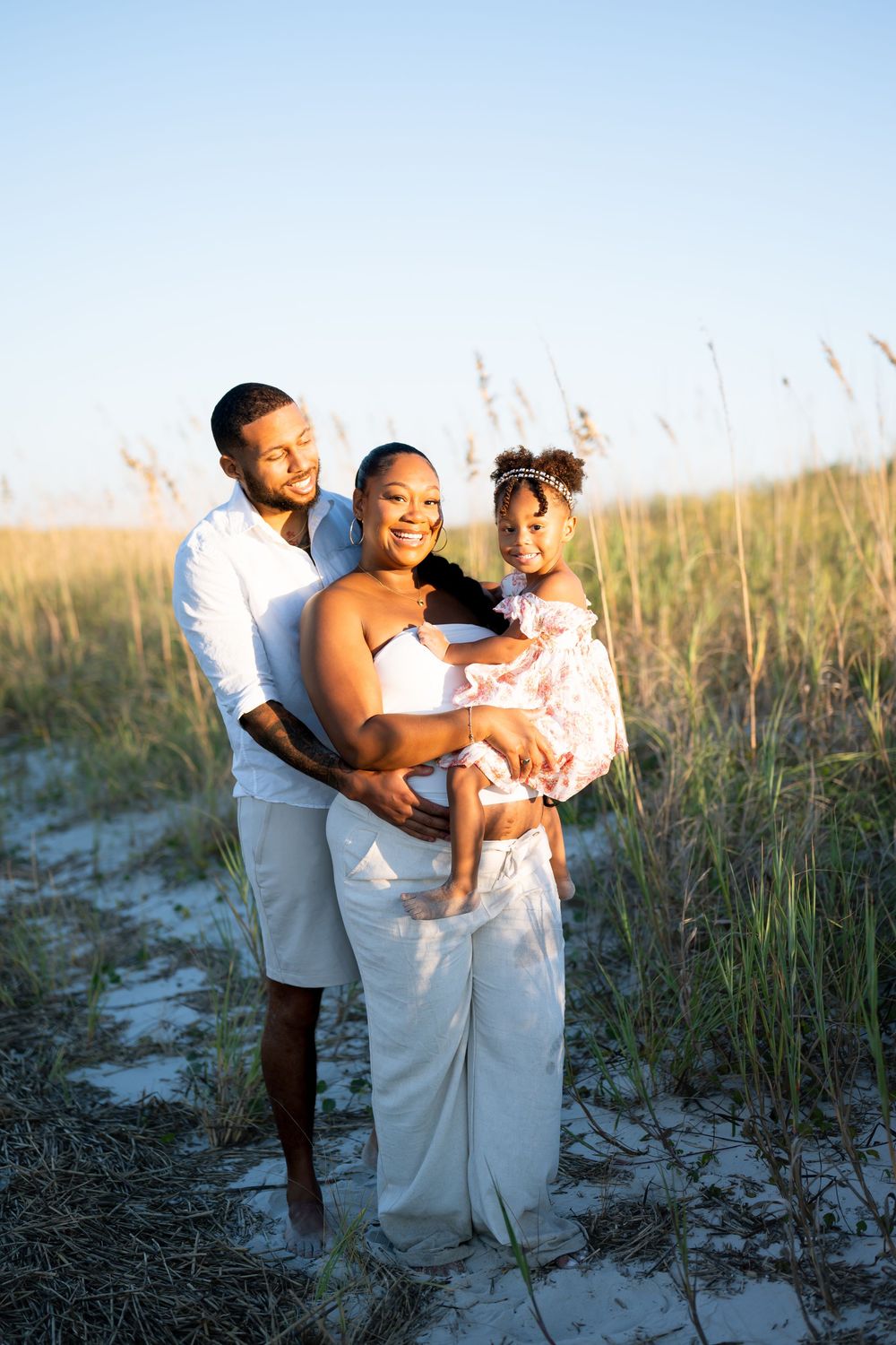 A loving family shares joyful moments together on a sandy beach with tall grass at sunset.
