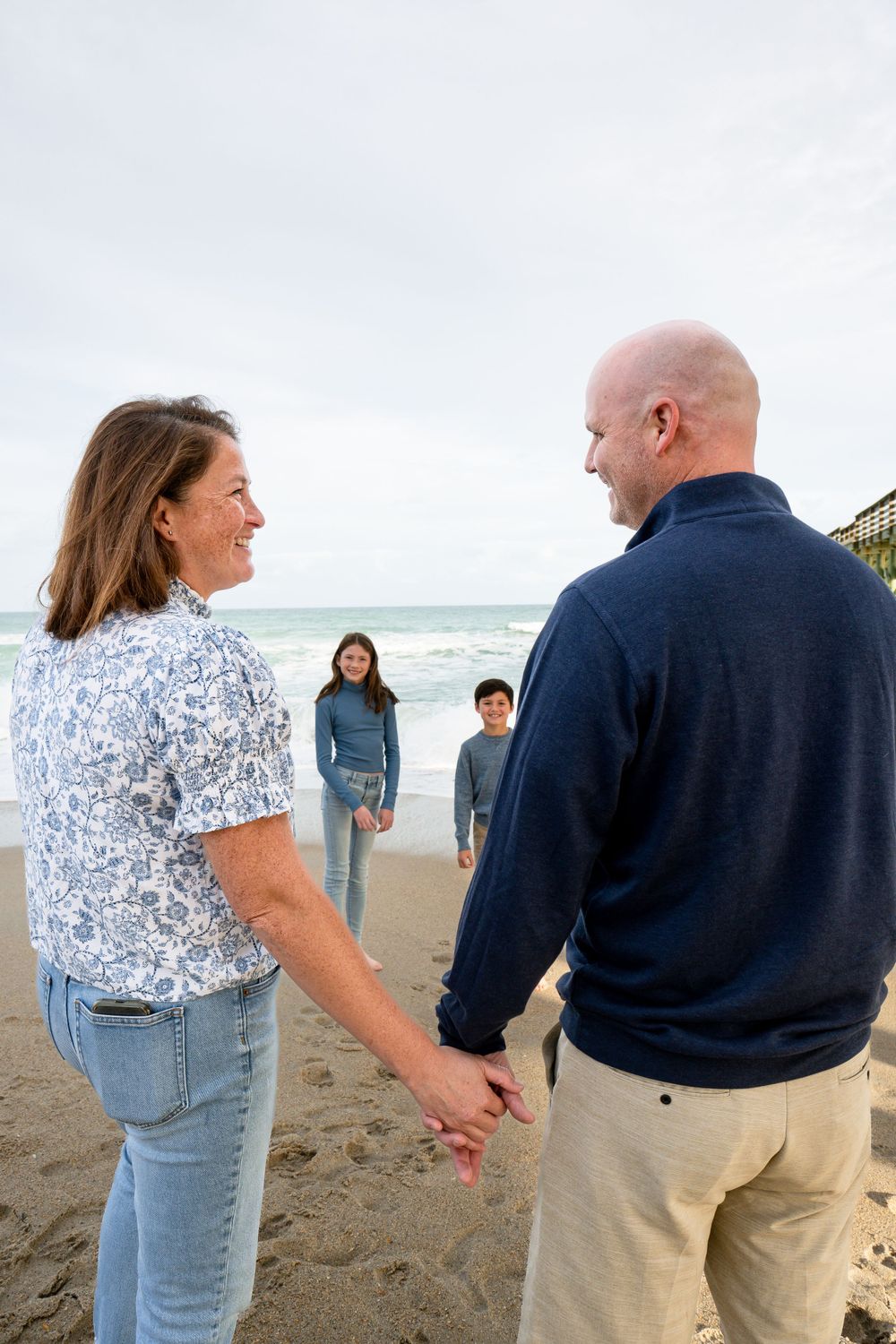 Couple holding hands on beach while two children stand in background near ocean.