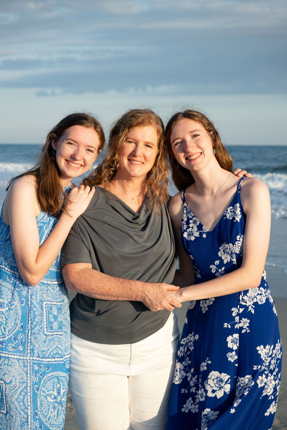 Three people in casual attire pose together on a beach at golden hour with ocean waves behind them.