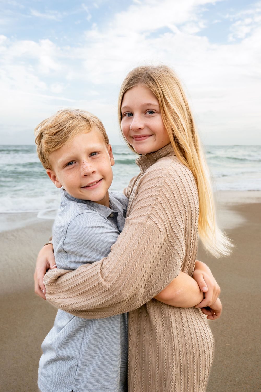 Two children hugging and smiling on beach with ocean waves in background.