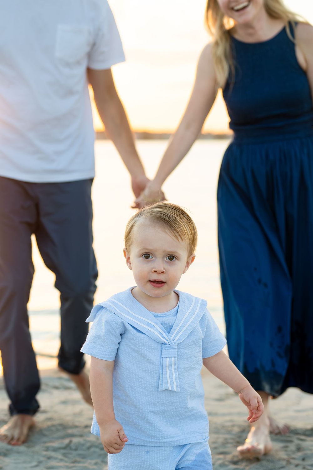 A small child in a light blue shirt walks on the beach while holding hands with adults in white and navy clothing.