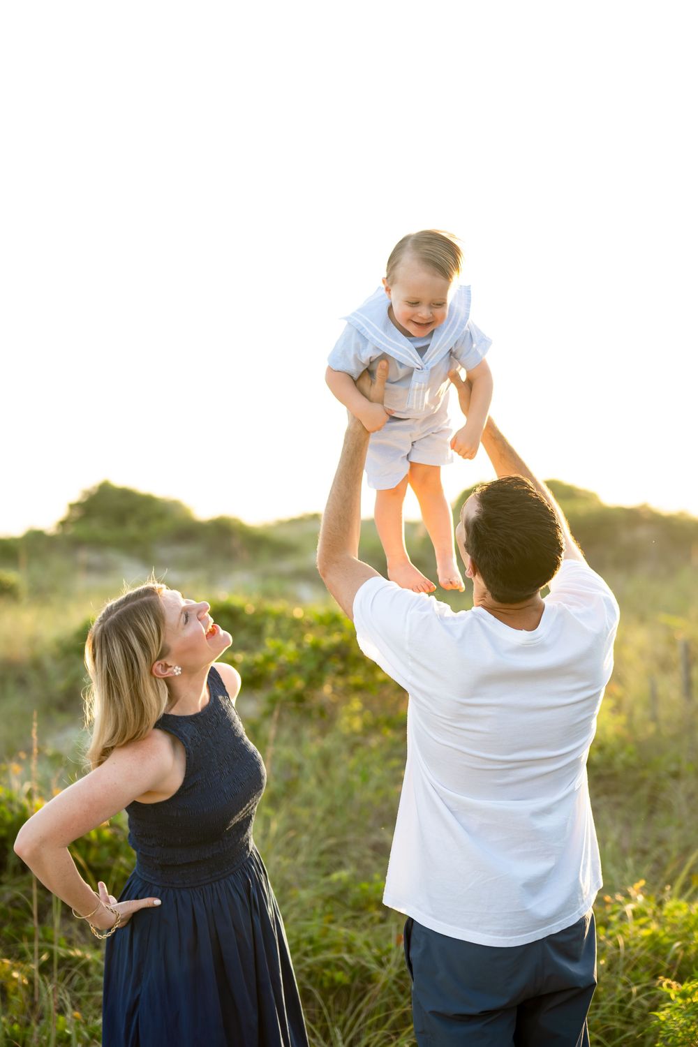 Parents playfully lift and swing their child in a sunny meadow during a summer evening.