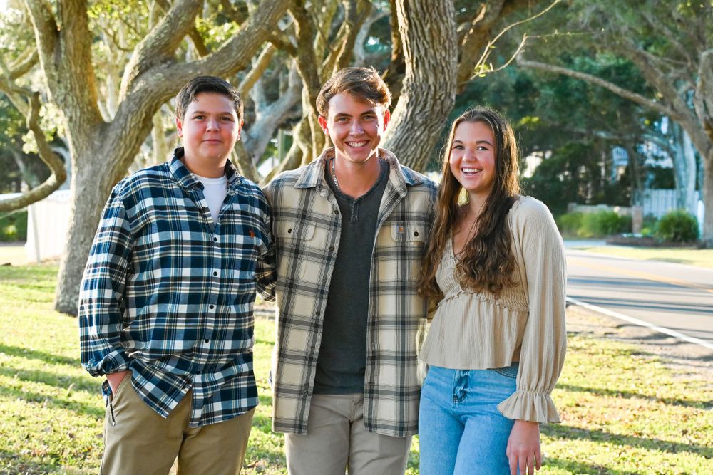 Three people in plaid shirts stand together outdoors under large oak trees.