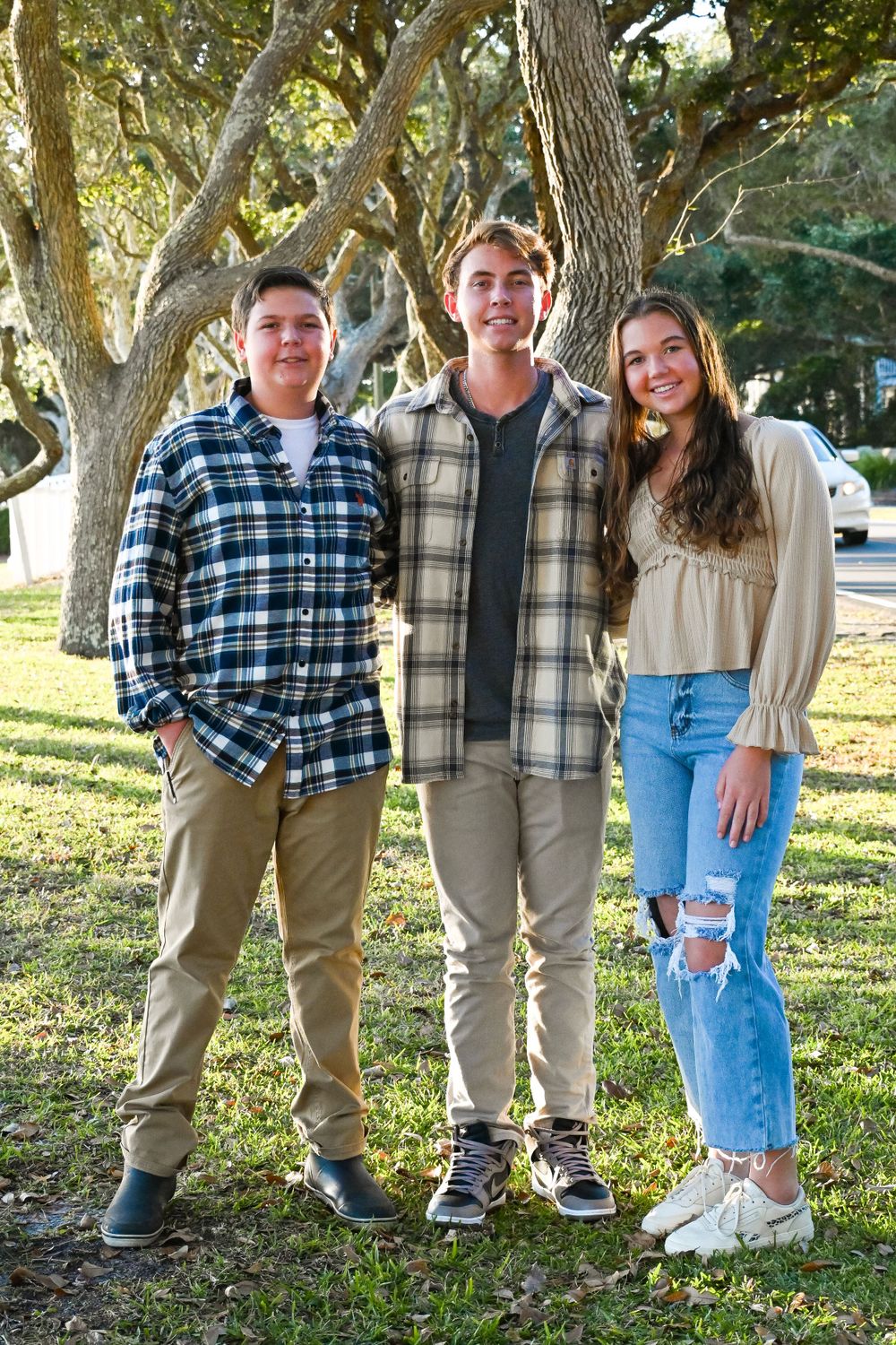 Three people in casual attire pose together under a canopy of oak trees on a sunny day.