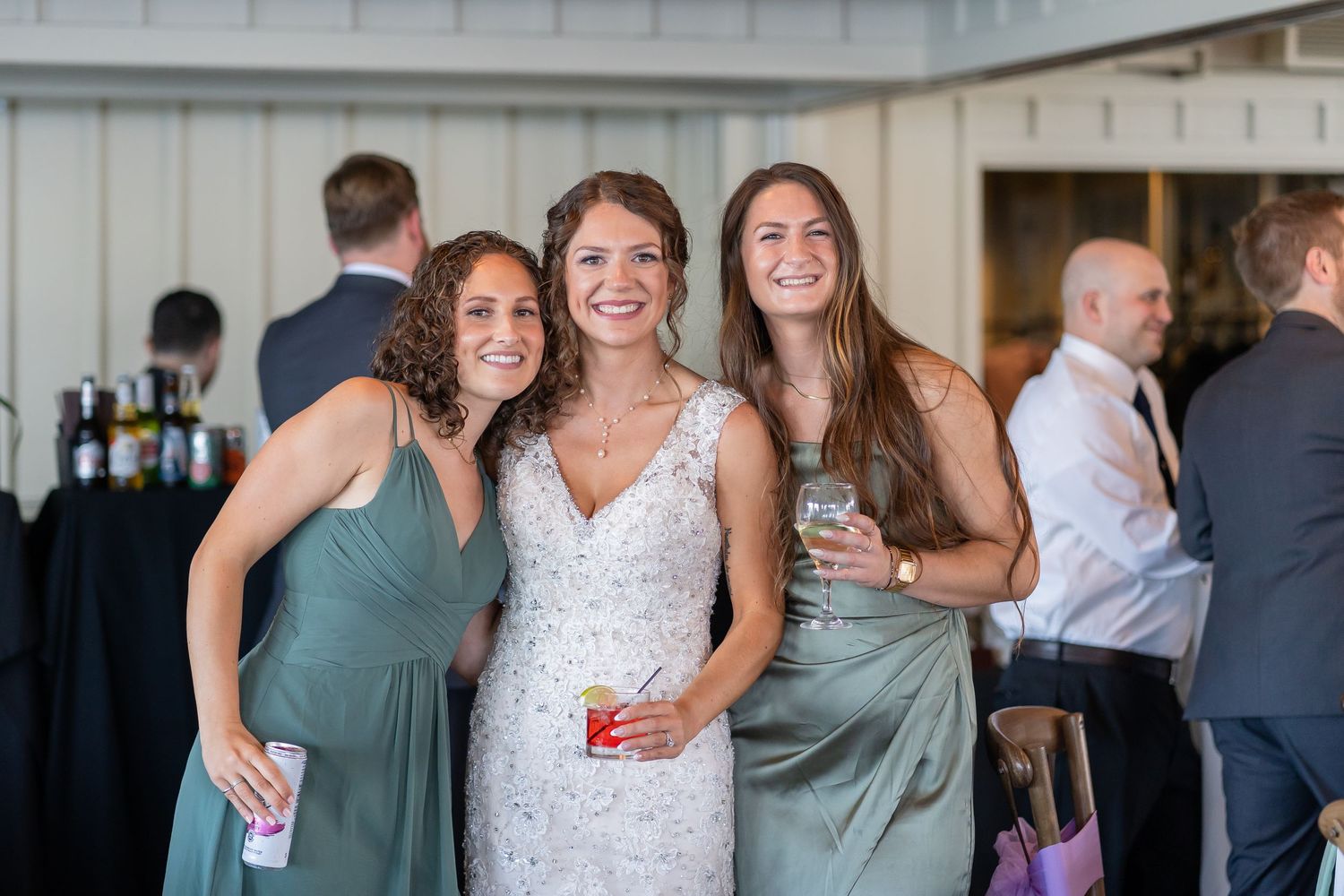 Three wedding attendees pose together at a reception, with two wearing sage green dresses and one in a white lace gown.