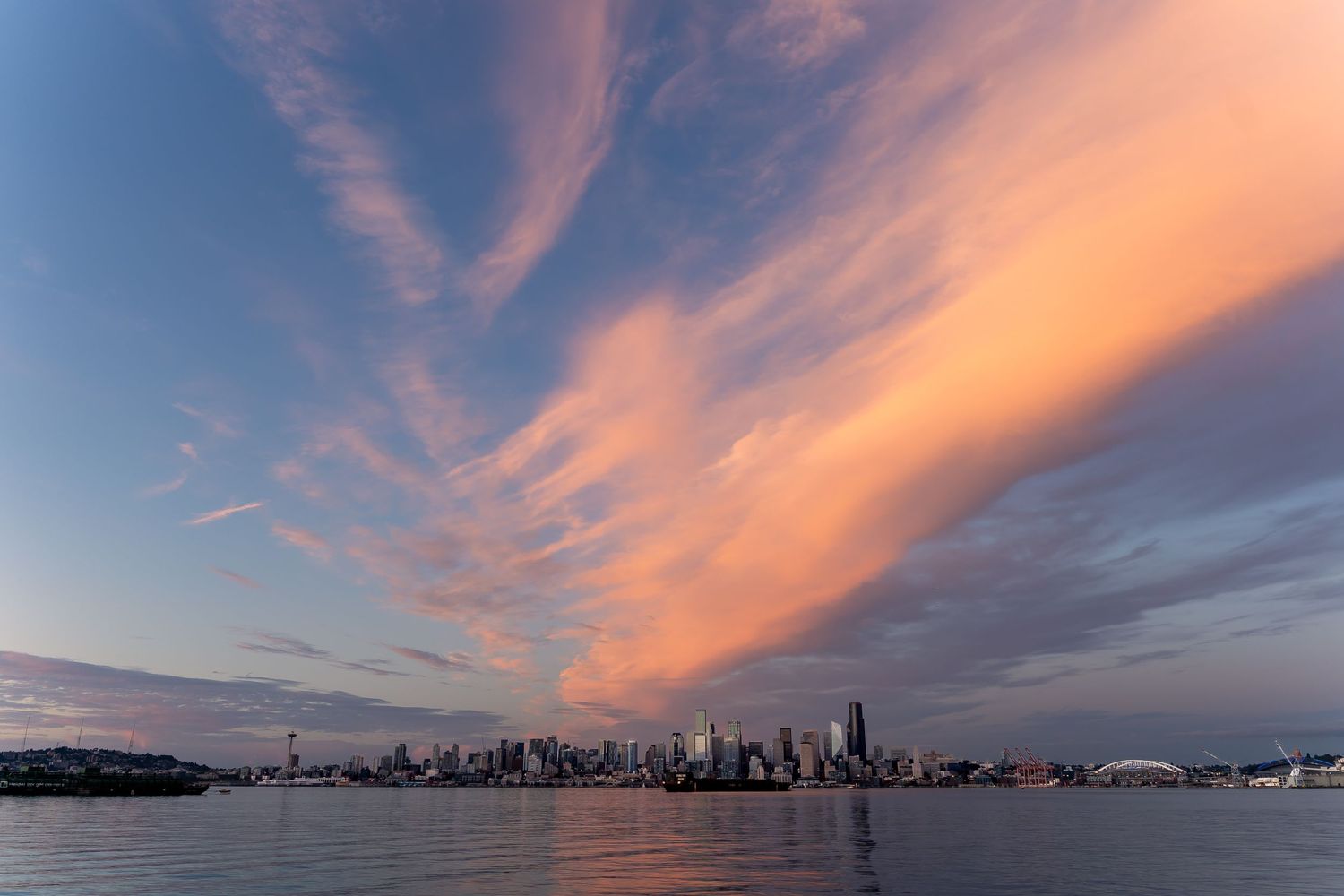 Pink and orange clouds sweep across the sky over the Seattle skyline reflecting in Elliott Bay at sunset.