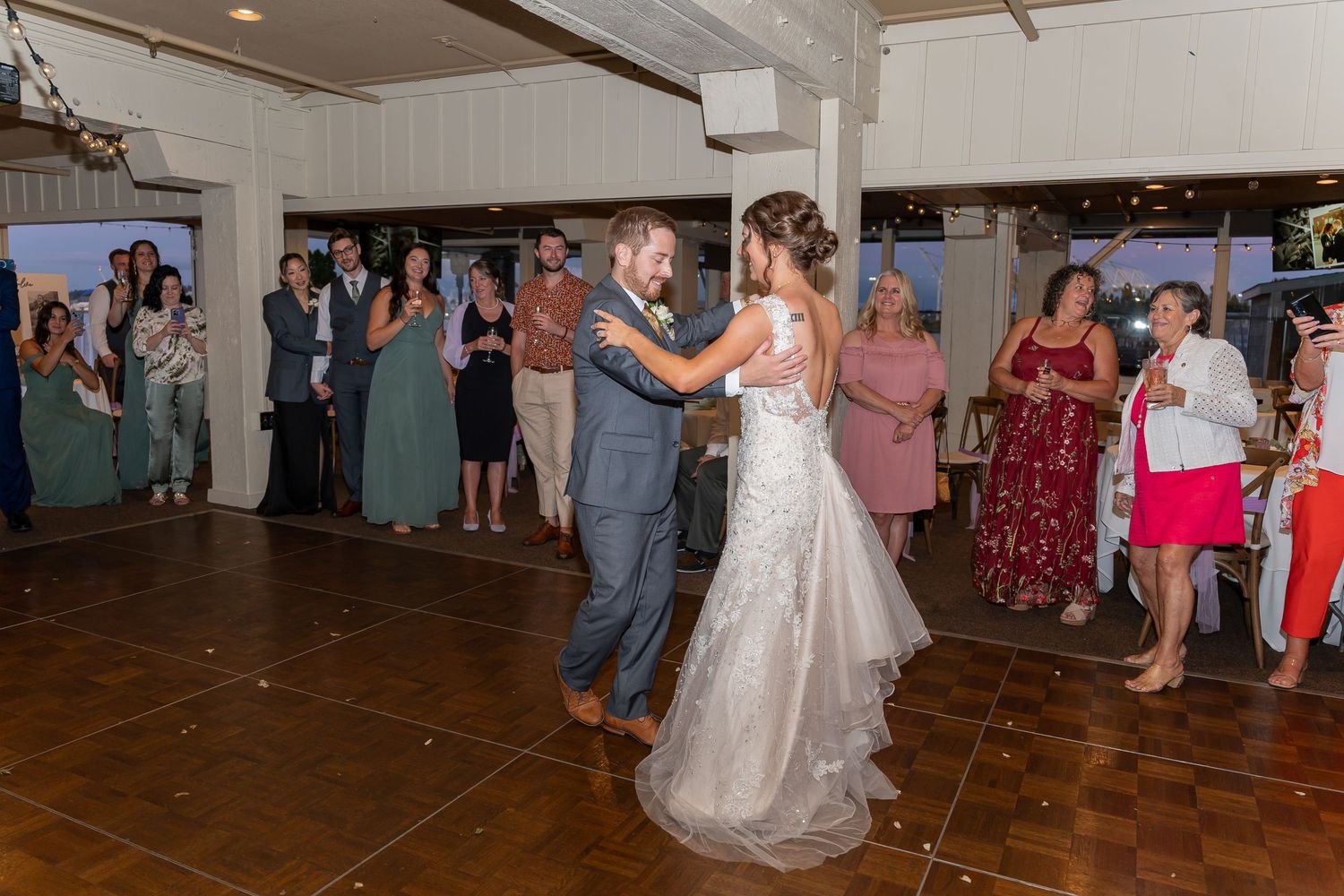 Wedding couple shares their first dance on a wooden dance floor while guests in teal bridesmaid dresses look on.