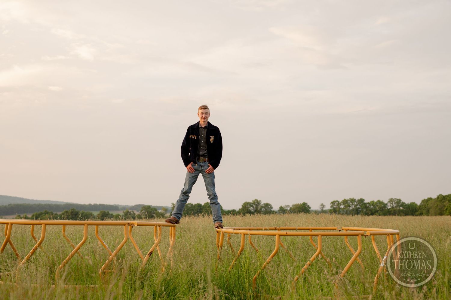Wide shot of student standing on wooden fence in rural field at sunset.