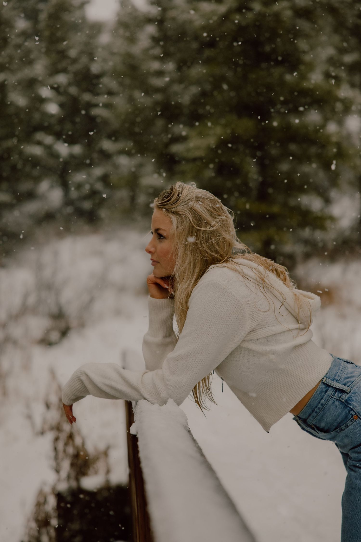 A person in a white sweater and jeans leans on a railing during a snowy winter day.