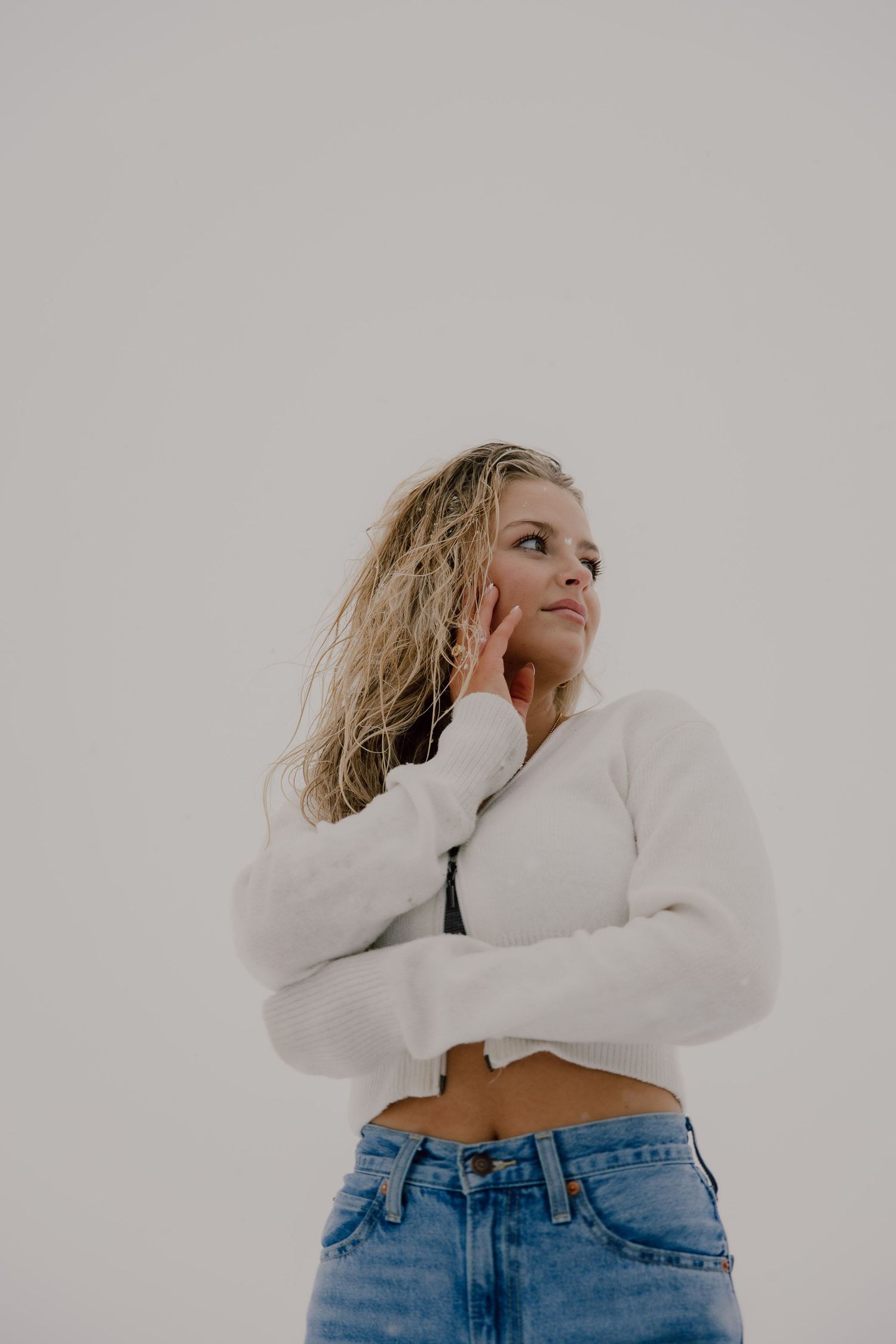 Person in white cropped sweater and blue jeans poses against light background with dreamy expression.