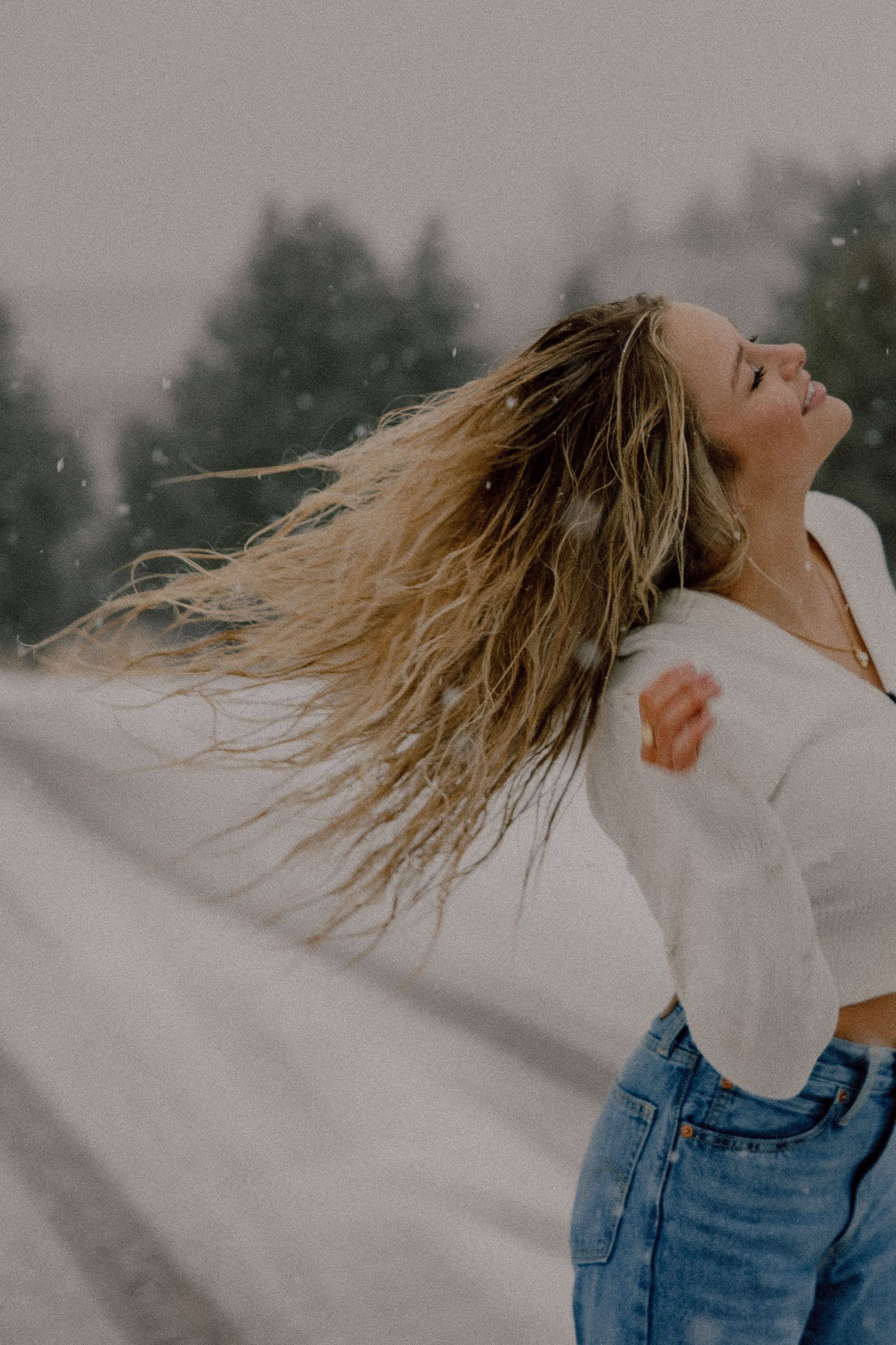 Person with windswept blonde hair wearing white sweater and jeans outdoors during snowy weather.
