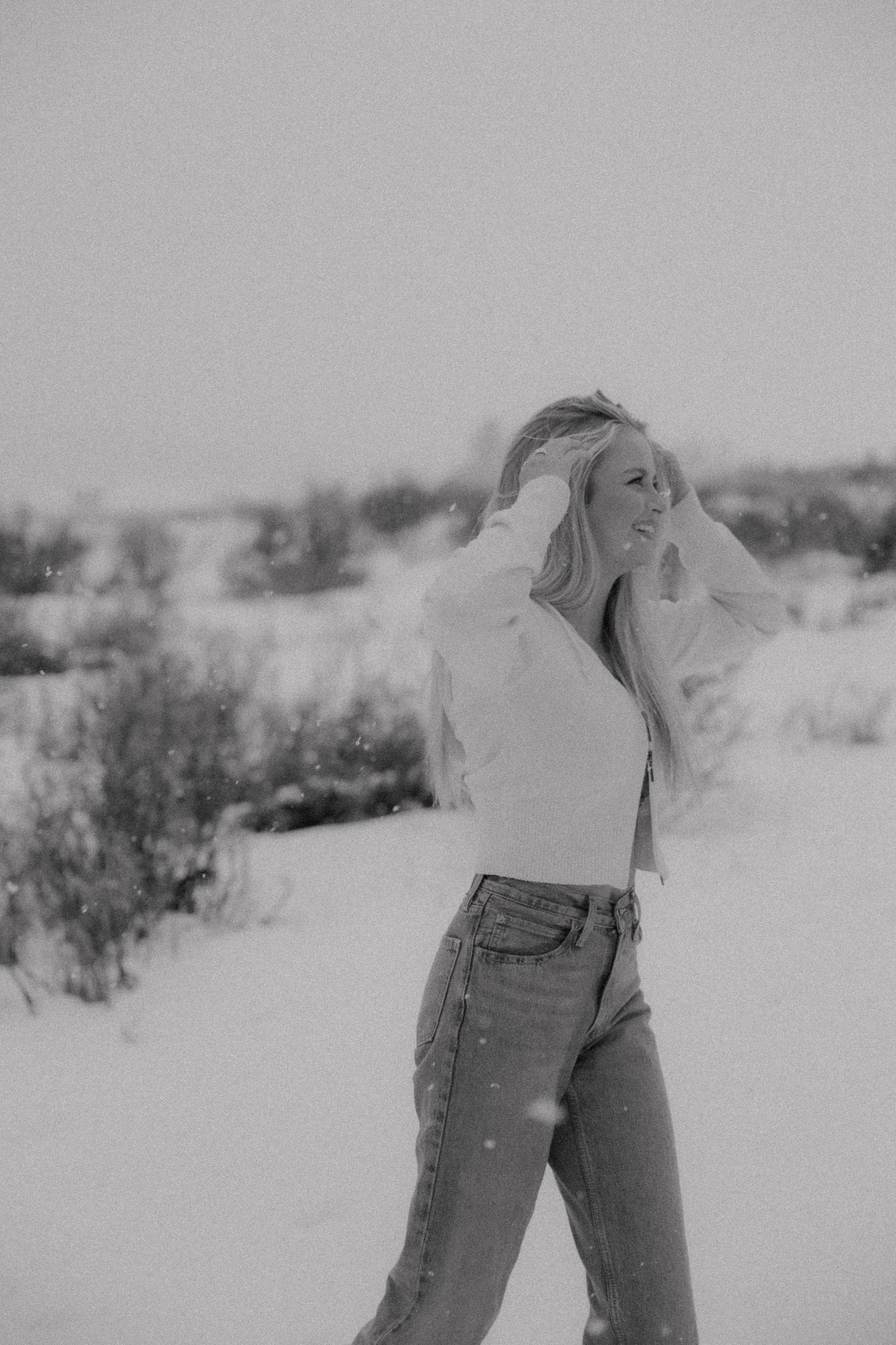 Black and white portrait of someone in blue jeans and white top posing in a snowy winter landscape.