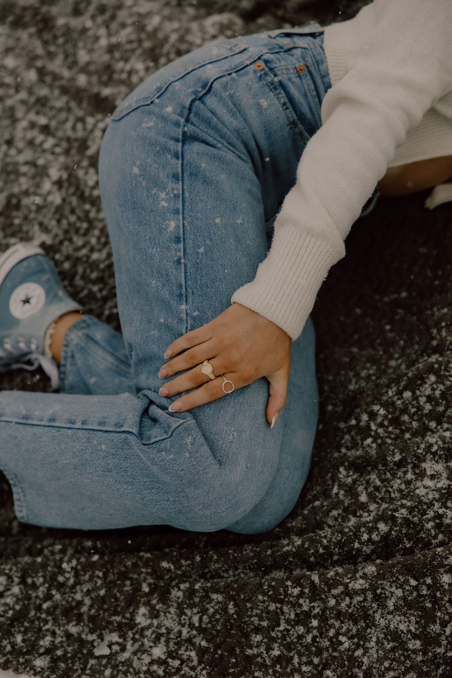 Close-up shot of someone wearing light blue jeans and a white sweater sitting casually on the ground with a ring on display.