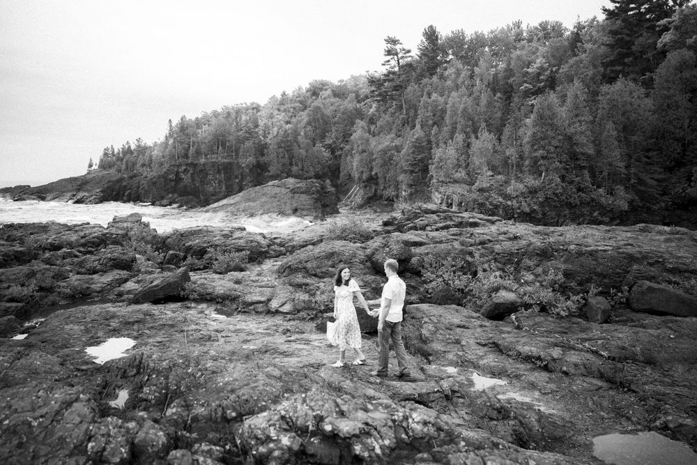 A couple in white outfits stands on rocky coastal cliffs with evergreen forest rising behind them in black and white.