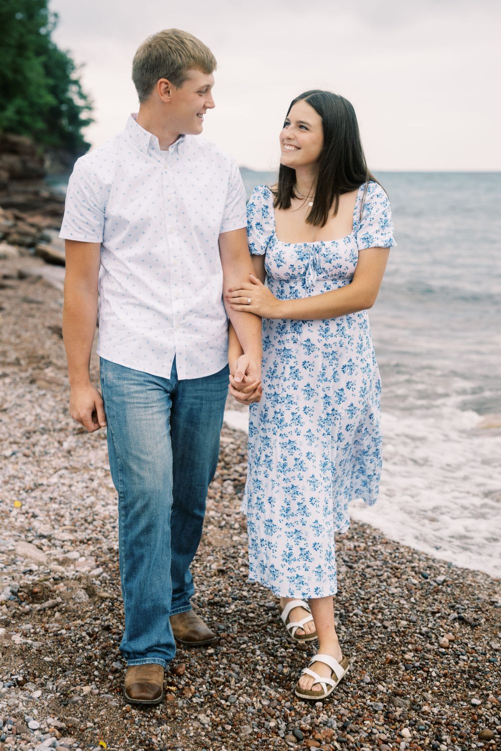 A couple in coordinated blue and white outfits walks hand in hand along a rocky beach shore.