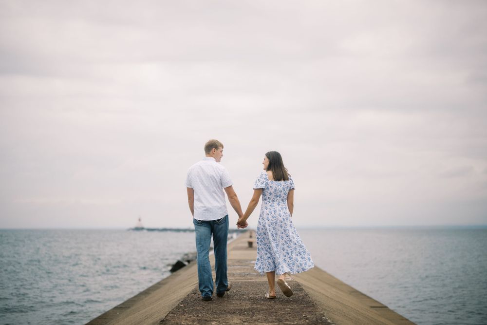 A couple holding hands walks together on a concrete pier extending into a misty ocean.